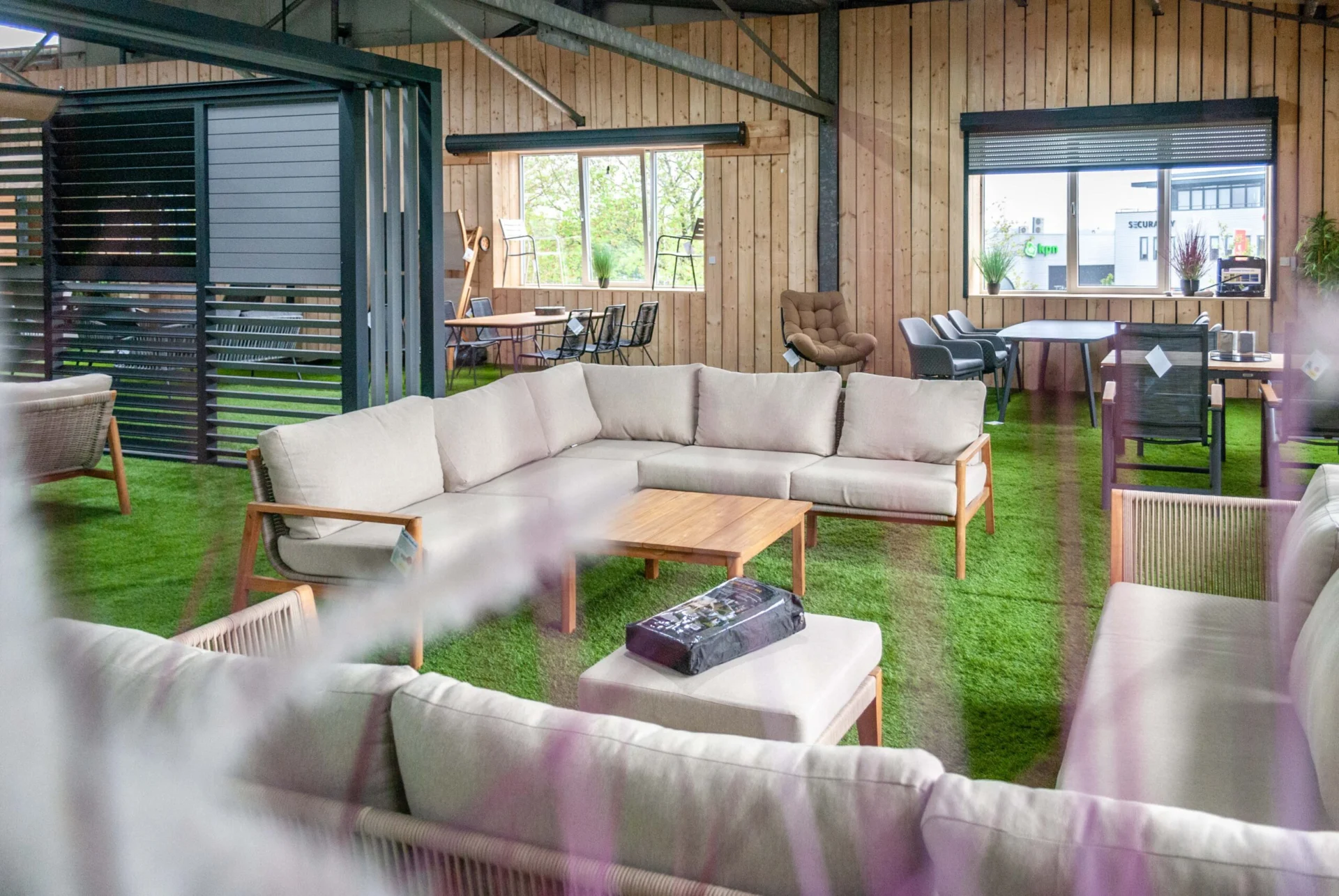 Een ruime loungeset met beige kussens en houten frame, geplaatst in een overkapping met houten wanden en kunstgras. Op de achtergrond staan verschillende tuinmeubels, tafels en stoelen.