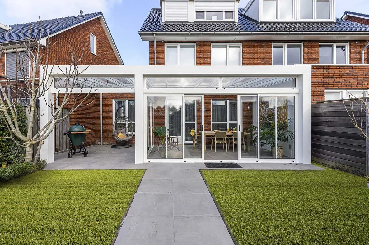 Een moderne witte overkapping met glazen schuifwanden, aangebouwd aan een rode bakstenen woning. De overkapping heeft een luxe inrichting met een houten eettafel, stoelen en planten, en grenst aan een tuin met kunstgras en grijze tuintegels.