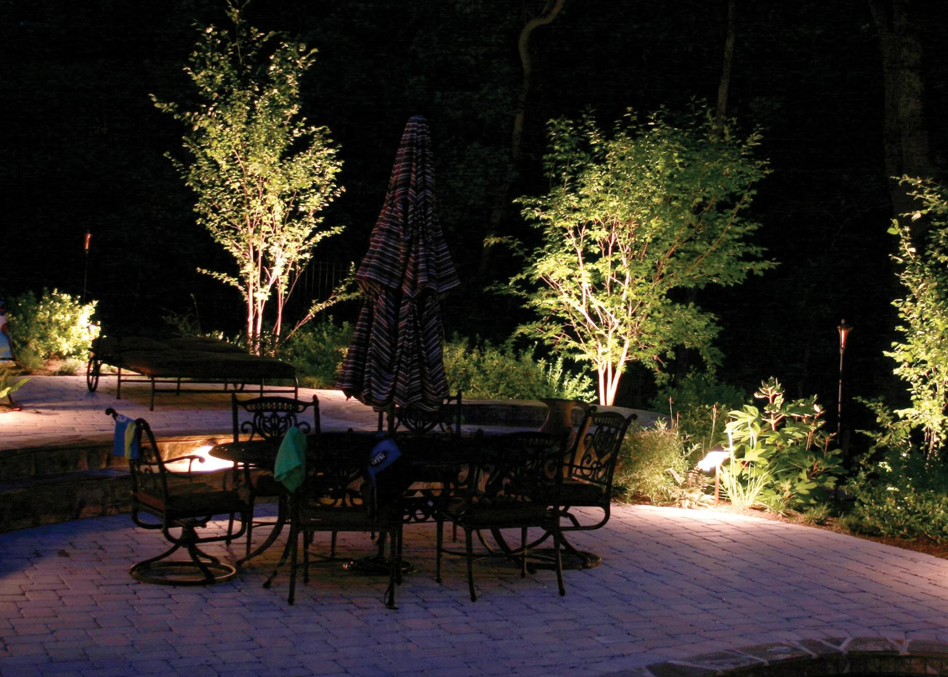 Verlichte tuin in de avond met sfeervolle spots die bomen, planten en een terras met metalen tuinmeubilair uitlichten.