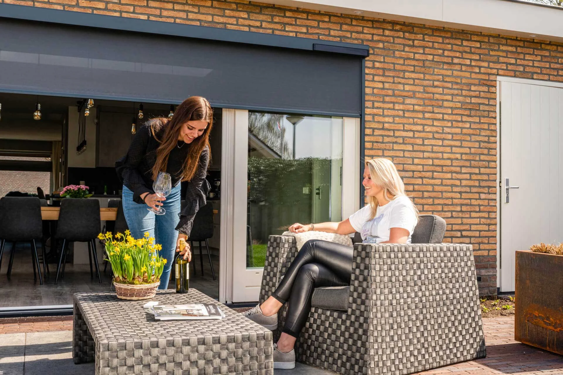 Donkergrijze screens gemonteerd op een bakstenen gevel, bieden effectieve zonwering en privacy voor een schuifpui met terras.