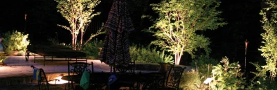 Verlichte tuin in de avond met sfeervolle spots die bomen, planten en een terras met metalen tuinmeubilair uitlichten.